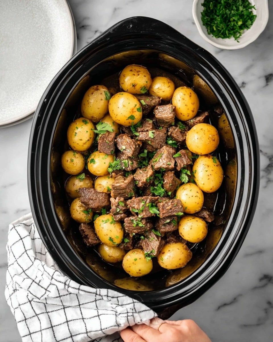 The image shows a black slow cooker filled with a simple meal. The bottom layer has many small round golden-brown potatoes spread evenly. On top of the potatoes is a layer of medium-sized dark brown chunks of cooked meat scattered throughout. Small pieces of chopped green parsley are sprinkled over the meat and potatoes for a touch of color. The slow cooker sits on a white marbled surface with part of a white marbled plate and a small white bowl with more parsley on the right side. A woman's hand is holding a white cloth with a black grid pattern on the left side. Photo taken with an iphone --ar 4:5 --v 7