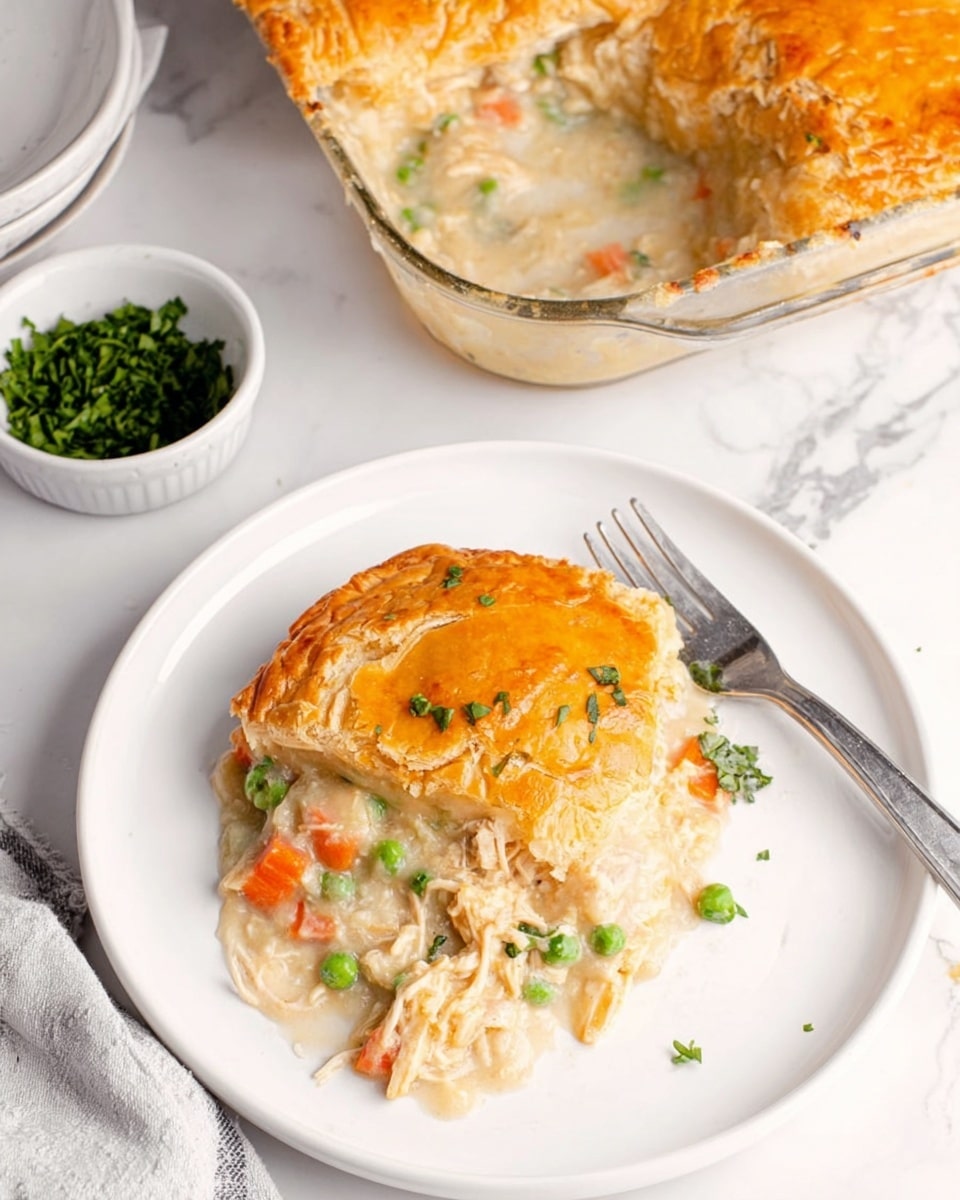 A round white plate shows a serving of chicken pot pie with a golden brown crust on top. The crust looks flaky and slightly cracked, revealing a creamy mixture underneath with visible pieces of white chicken, orange carrots, and green peas. The creamy filling has a light color with small vegetable bits scattered around. Next to the plate, a silver fork rests partly on the plate. In the background, there is a glass baking dish with some of the pot pie missing, showing the same creamy white and orange filling. A small white bowl filled with chopped green herbs sits nearby on a white marbled surface. photo taken with an iphone --ar 4:5 --v 7