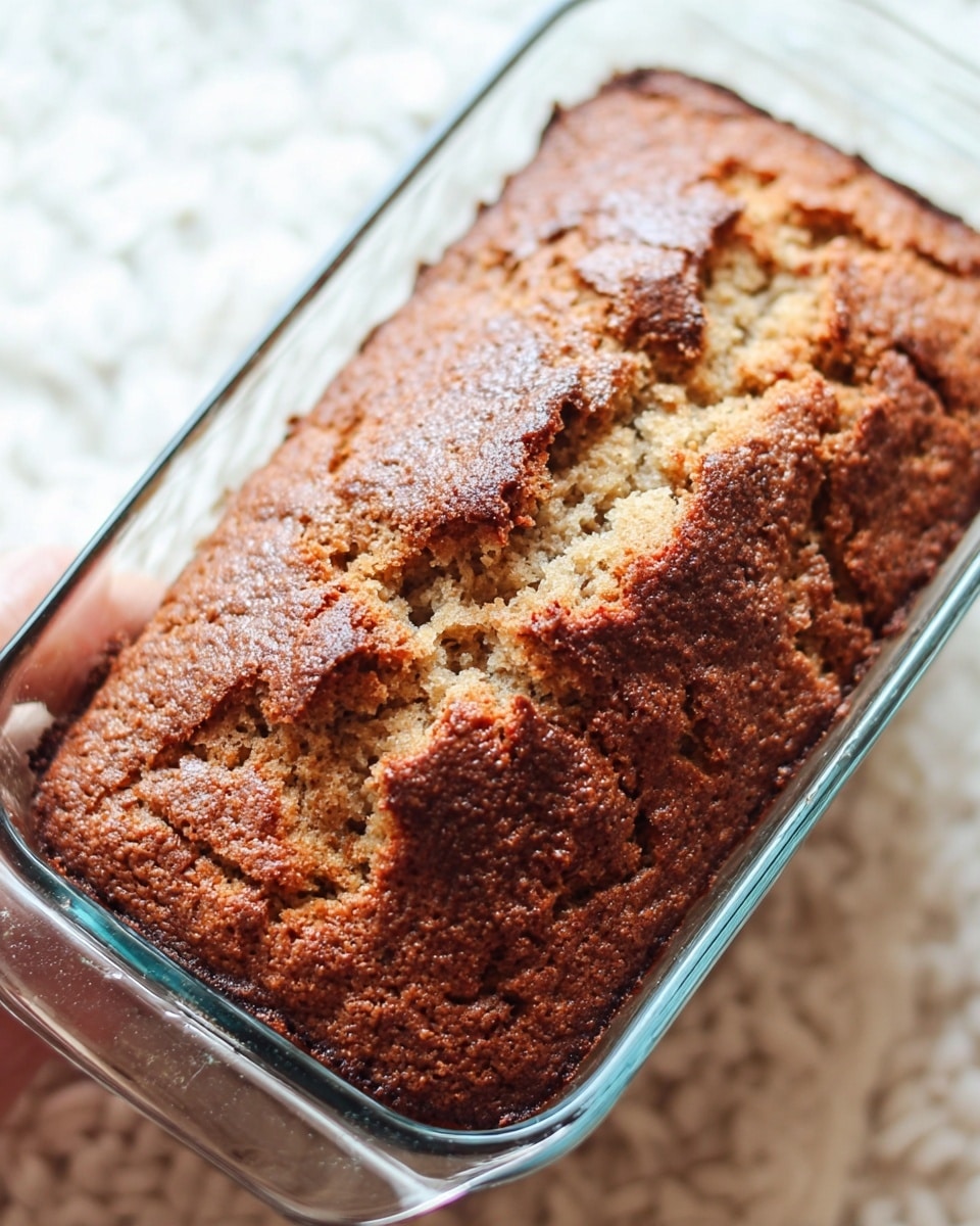 A close-up image of a baked loaf in a clear glass rectangular dish, showing a rough, golden brown crust with darker brown patches and cracked, uneven texture on the top layer. The loaf appears to be well risen with a slightly crispy surface. The background features a white marbled texture, and a woman's hand is faintly visible holding the dish on the right side. Photo taken with an iphone --ar 4:5 --v 7