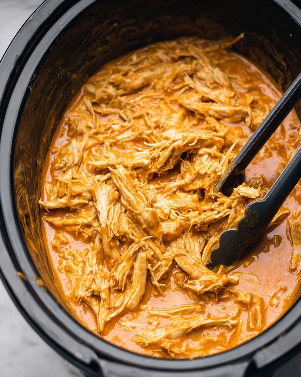 The image shows a black slow cooker filled with shredded chicken mixed in a thick, creamy orange sauce. The chicken is pulled into thin strands and is fully coated with the sauce that has a smooth and slightly glossy texture. There are black tongs stirring the chicken in the pot, with one side of the tongs visible on the right and the other on the upper side. The background surface is white marbled, adding a clean look to the image. photo taken with an iphone --ar 4:5 --v 7