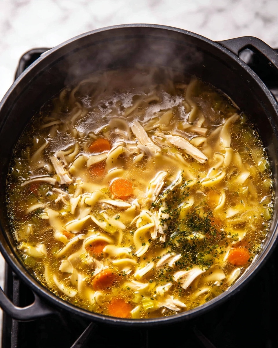 A close-up of a pot filled with thick chicken noodle soup. The soup has three main layers: a clear golden broth with oil glistening on the surface, pieces of shredded pale chicken and small chunks of orange carrots and green celery floating throughout, and short, flat egg noodles spreading inside. There is also a visible layer of dark green herbs sprinkled in the middle, adding texture and depth. Steam is rising from the soup, showing it is hot and freshly cooked. The pot is black and placed on a stove with a white marbled surface in the background. photo taken with an iphone --ar 4:5 --v 7