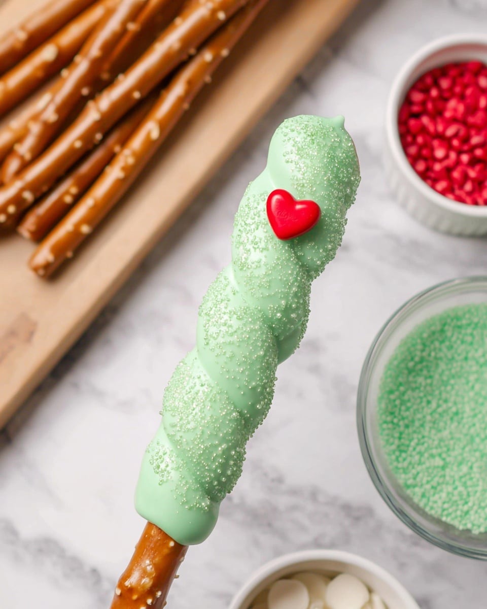 A single twisted pretzel stick coated with a thick layer of shiny, light green candy, sprinkled with fine sugar crystals for texture. On the front middle part of the candy layer, there is a small, bright red heart-shaped candy decoration. The pretzel stick shows at the bottom with its golden brown color and salt crystals. The background features a white marbled surface with a wooden board holding more plain pretzel sticks on the left, two small white bowls at the top right filled with green sugar crystals and red heart-shaped candies, and a glass bowl filled with the same light green candy coating on the right side. photo taken with an iphone --ar 4:5 --v 7