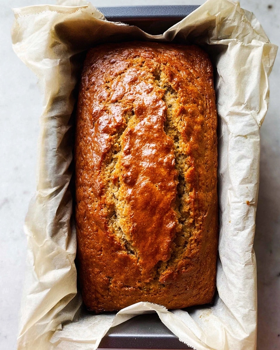 A rectangular loaf of golden-brown banana bread with a shiny, slightly cracked top sits inside a dark baking pan lined with wrinkled white parchment paper, the loaf showing a rough texture indicating moisture and soft crumb inside, all placed on a white marbled surface photo taken with an iphone --ar 4:5 --v 7