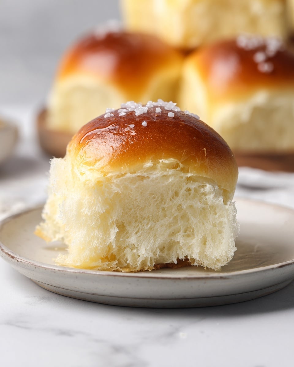 A soft, shiny golden brown roll with a smooth top is placed in the center of a round white plate. The roll is split open, showing its fluffy, light cream-colored inside with a moist texture. The top of the roll is sprinkled with a few coarse white salt crystals. In the blurred background, several more rolls appear stacked together. The scene is set on a white marbled surface. photo taken with an iphone --ar 4:5 --v 7