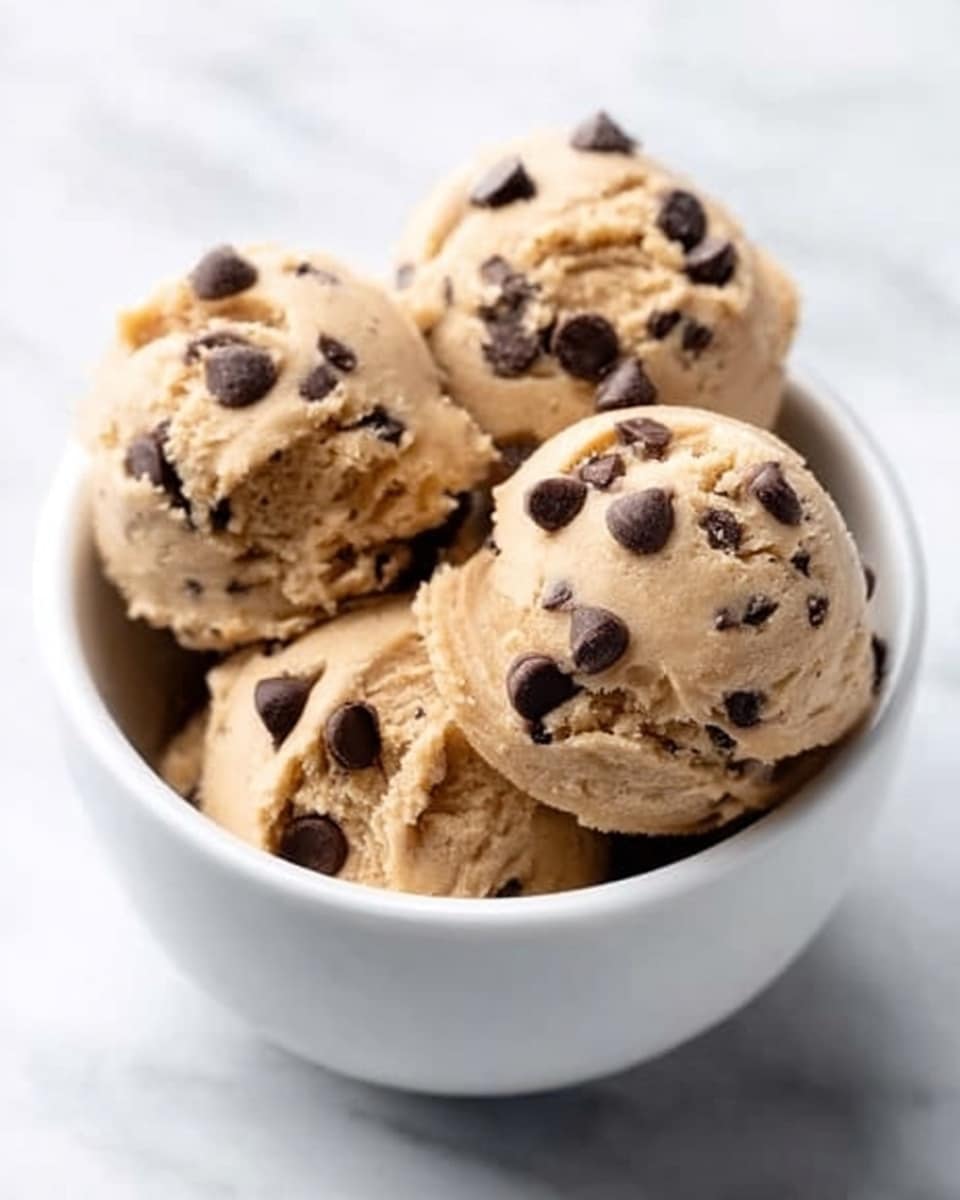 A white bowl filled with four large scoops of light brown cookie dough, each scoop dotted generously with dark brown chocolate chips. The cookie dough looks soft and slightly rough in texture, with the chocolate chips embedded throughout, some resting on the surface for a rich contrast. The bowl sits on a white marbled surface that adds a clean and bright background to the image. photo taken with an iphone --ar 4:5 --v 7