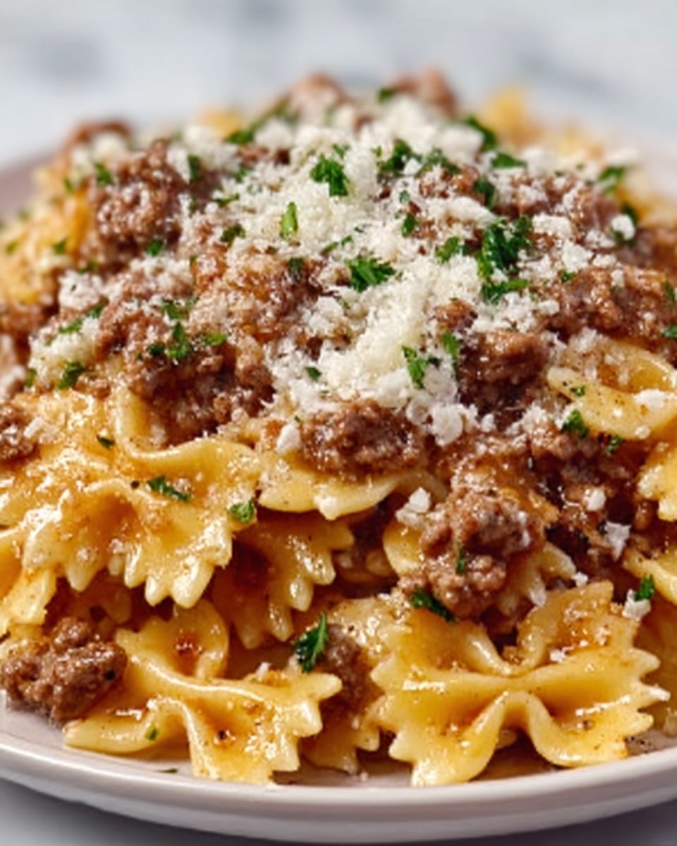 This image shows a close-up of a dish with three clear layers: the bottom layer is light orange bowtie pasta with a shiny, slightly soft texture; the middle layer is made of small chunks of browned ground meat evenly spread among the pasta; the top layer has finely grated white cheese scattered over the meat and pasta, with small bits of green herbs sprinkled as garnish. The dish is served in a shallow, white plate on a white marbled surface. Photo taken with an iphone --ar 4:5 --v 7