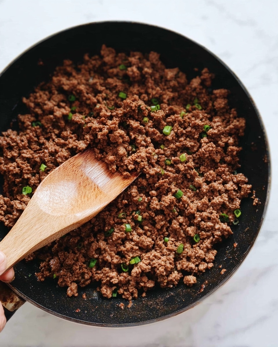 A black pan filled with cooked ground meat mixed with small green onion pieces scattered across the meat, creating small green spots on the brown texture. A wooden spoon is placed inside the pan, resting on the cooked meat, and a woman's hand is holding the handle of the spoon. The surface under the pan is white marbled texture. photo taken with an iphone --ar 4:5 --v 7