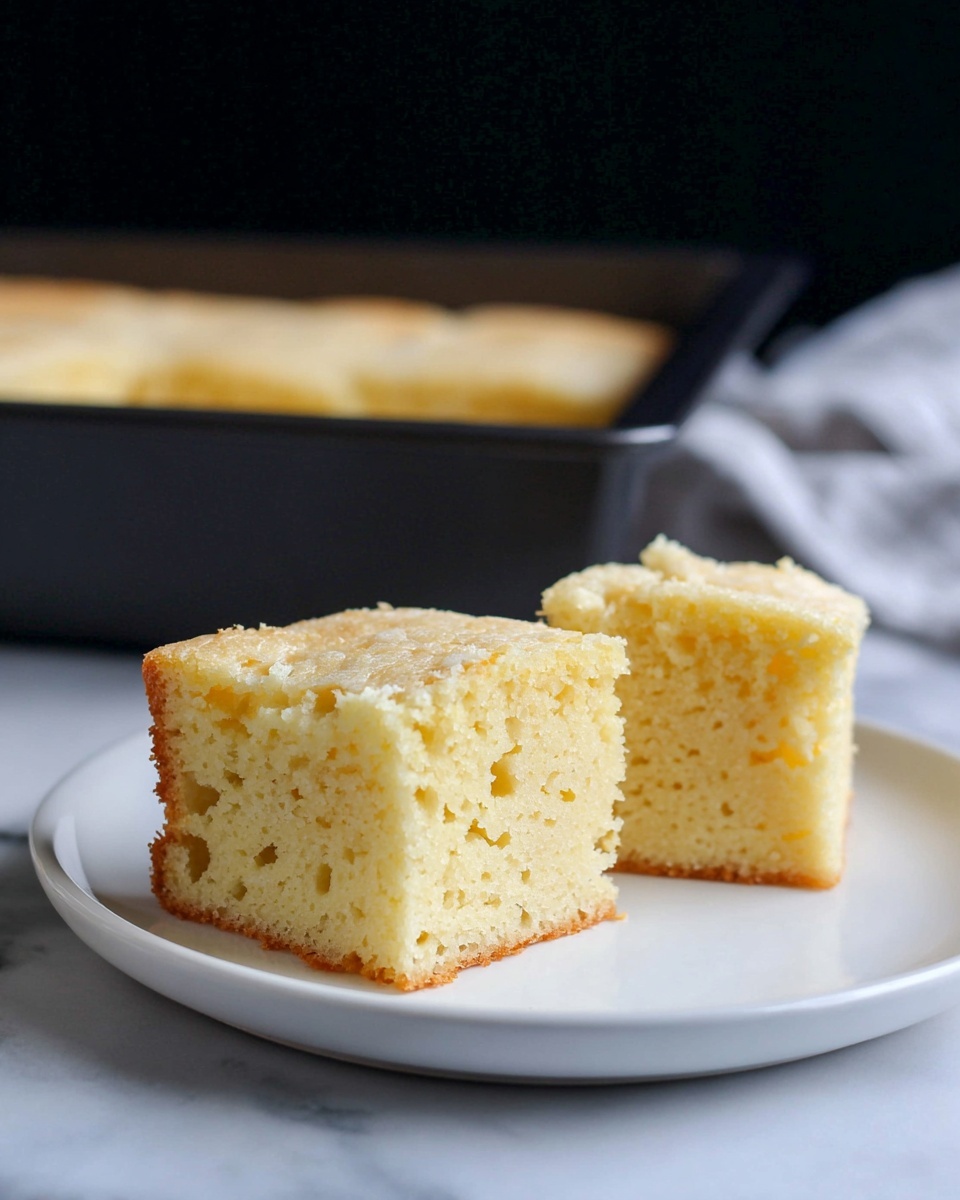 Two square pieces of light yellow cake with a soft, moist texture and small holes throughout, sitting side by side on a white plate. The cake has a thin, slightly crumbly pale crust on the top and edges. Behind the plate, there is a black baking pan filled with more of the same cake. The scene is set on a white marbled surface with a blurred dark background. photo taken with an iphone --ar 4:5 --v 7