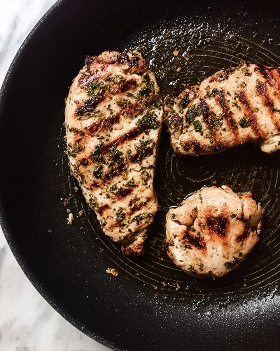The image shows three cooked pieces of meat in a black frying pan with a slightly shiny, oily surface. The meat pieces are light brown with visible grill marks and dark brown charred spots; they have a speckled layer of green herbs and spices covering their top surfaces. The largest piece is curved and positioned vertically on the left side, the second piece is partially shown at the top right, and the smallest rounded piece is at the bottom right. The pan's surface is textured with circular ridges, and there are small pools of oil and bits of browned seasoning around the meat. The whole scene is set on a white marbled surface. Photo taken with an iphone --ar 4:5 --v 7