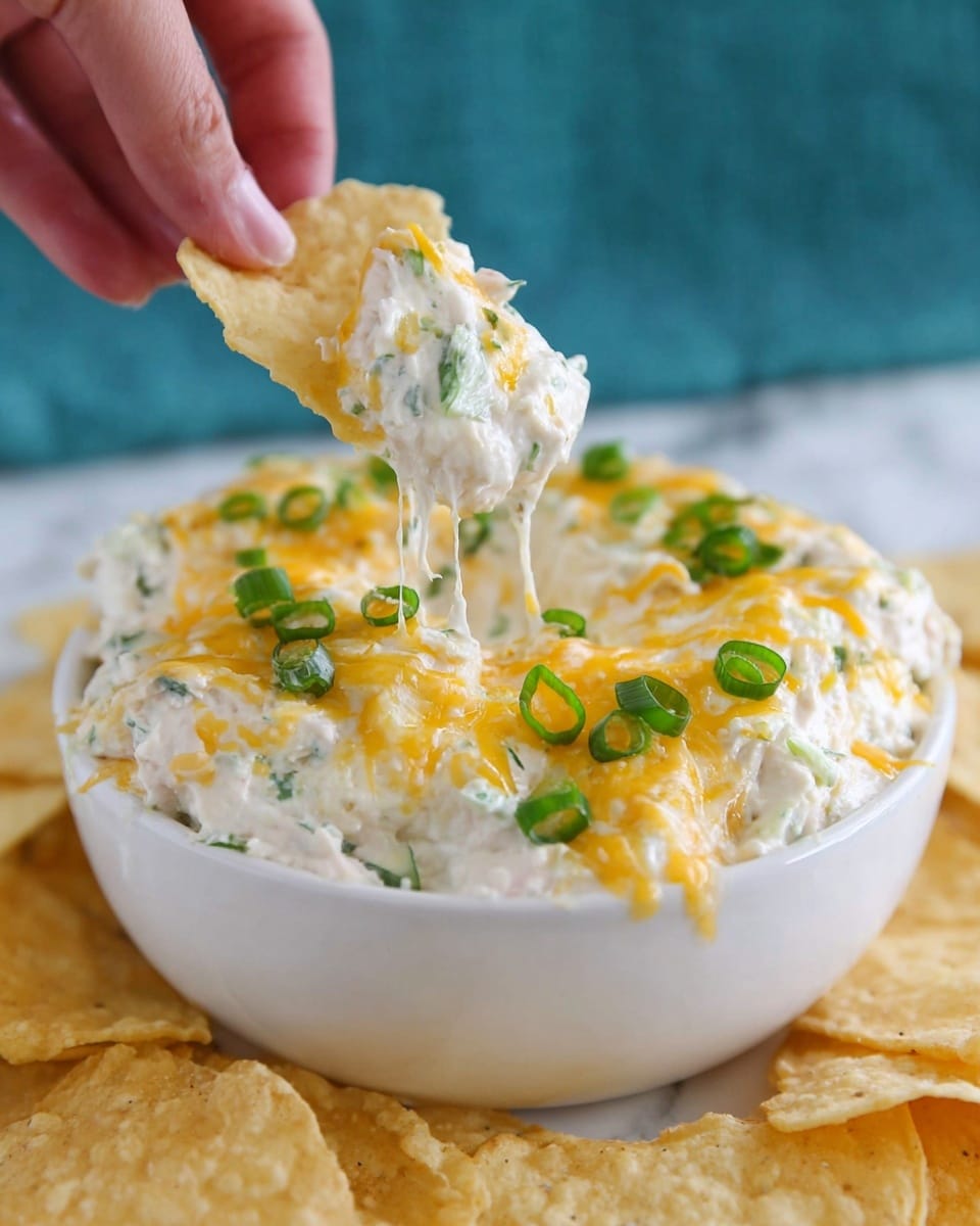 A white bowl filled with a creamy dip that has a soft white color as the base layer, mixed with small bits of green herbs. On top, there is a layer of melted yellow cheese scattered unevenly. Green slices of spring onion are spread over the cheese, adding a fresh look. A woman's hand is holding a light yellow chip being dipped into the creamy mixture, pulling up some cheese and dip that stretches between the bowl and chip. The background shows more chips on a white marbled surface with a slight blue tint out of focus. Photo taken with an iphone --ar 4:5 --v 7