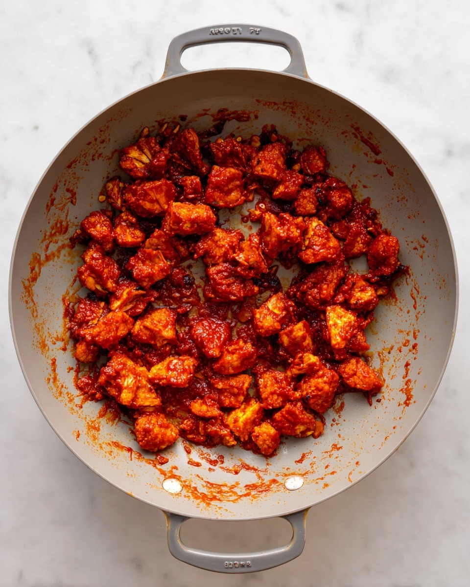 Inside a light gray pan with two handles, there are many small, uneven pieces of food coated in a thick, bright red sauce. The pieces are spread mostly on one side of the pan, showing a glossy and sticky texture. The pan sits on a white marbled surface, and some sauce spots are scattered around the pan's inner sides. photo taken with an iphone --ar 4:5 --v 7