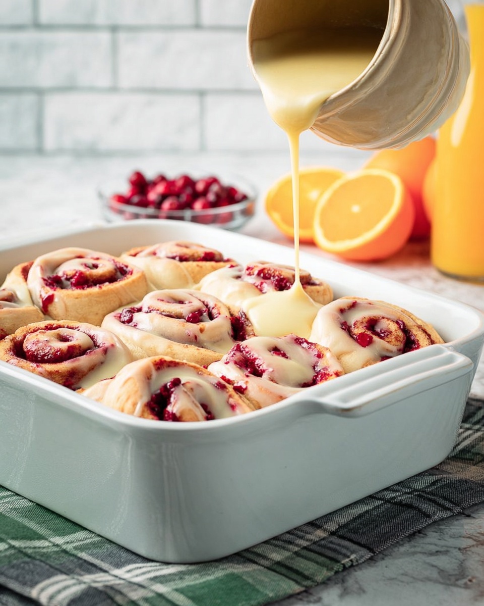 A white baking dish filled with nine cinnamon rolls swirled with red berry filling, arranged in three rows of three. A thick light yellow icing is being poured from a beige jug in the top left corner onto the center roll, creating a creamy layer over the red swirls and soft dough. The baking dish sits on a gray and white checked cloth atop a white marbled surface. In the background, there are whole and sliced oranges, some cranberries, and a bottle of orange juice against a white brick wall. photo taken with an iphone --ar 4:5 --v 7