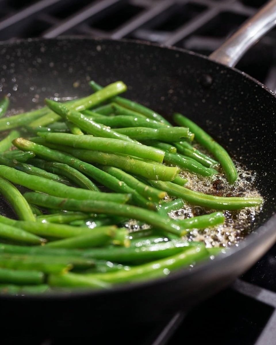A close-up of fresh green beans being cooked in a black, speckled pan with some bubbling oil around them. The green beans are bright and shiny, laying mostly flat and overlapping each other inside the pan. The pan is on a stove with dark grates barely visible in the background, and the overall focus is on the green beans and the sizzling oil. photo taken with an iphone --ar 4:5 --v 7