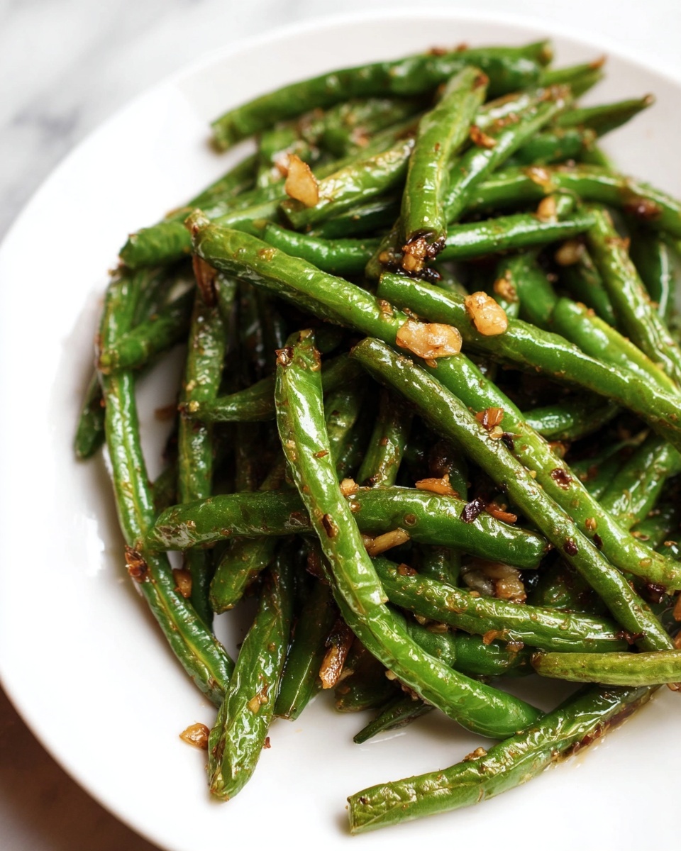 A close-up view of a white plate filled with cooked green beans, showing about one layer of beans piled loosely in the center. The green beans are glossy with a slightly wrinkled texture and some darker browned spots, coated with small pieces of cooked garlic scattered throughout. The plate sits on a white marbled surface. Photo taken with an iphone --ar 4:5 --v 7