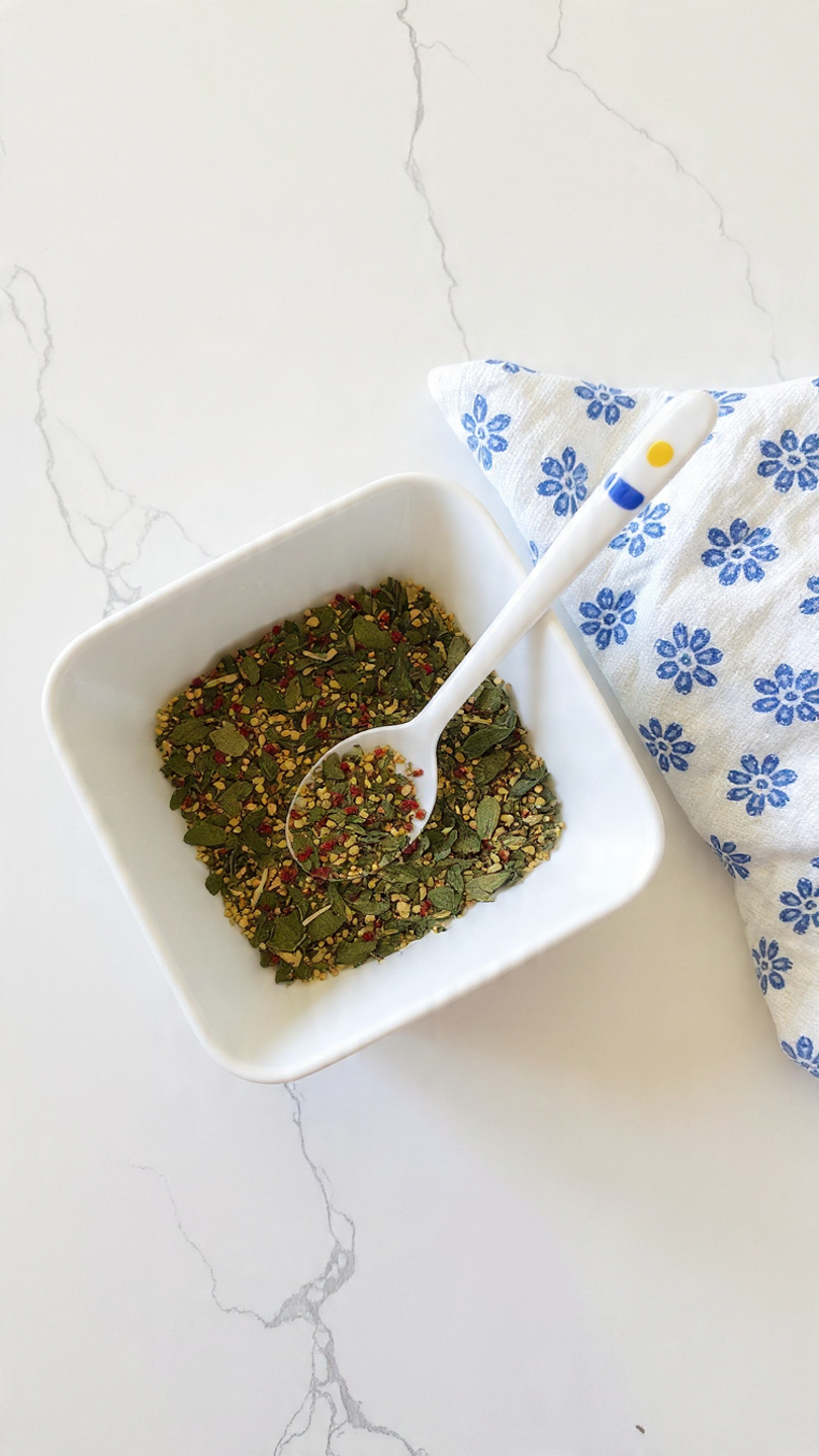 The image shows a white square bowl filled with a colorful mix of dried herbs and spices including small green leaves, red pepper flakes, and some yellow seeds. Inside the bowl, there is a small spoon with a white handle decorated with blue and yellow lines. The bowl is placed on a white marbled surface, and next to it is a white cloth with a blue flower pattern. Photo taken with an iphone --ar 4:5 --v 7