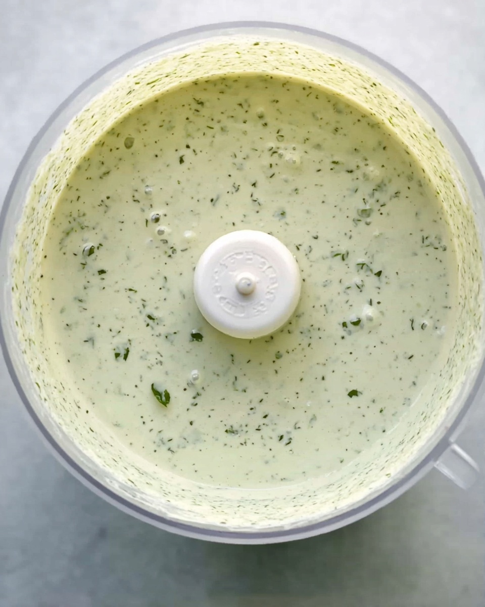 The image shows a close-up view of a thick, light green sauce inside a clear round food processor bowl. The sauce has a creamy texture with many small air bubbles scattered on its surface, giving it a slightly frothy look. In the center, there is a beige plastic blade attachment partially covered in the sauce. The background is a white marbled texture. photo taken with an iphone --ar 4:5 --v 7