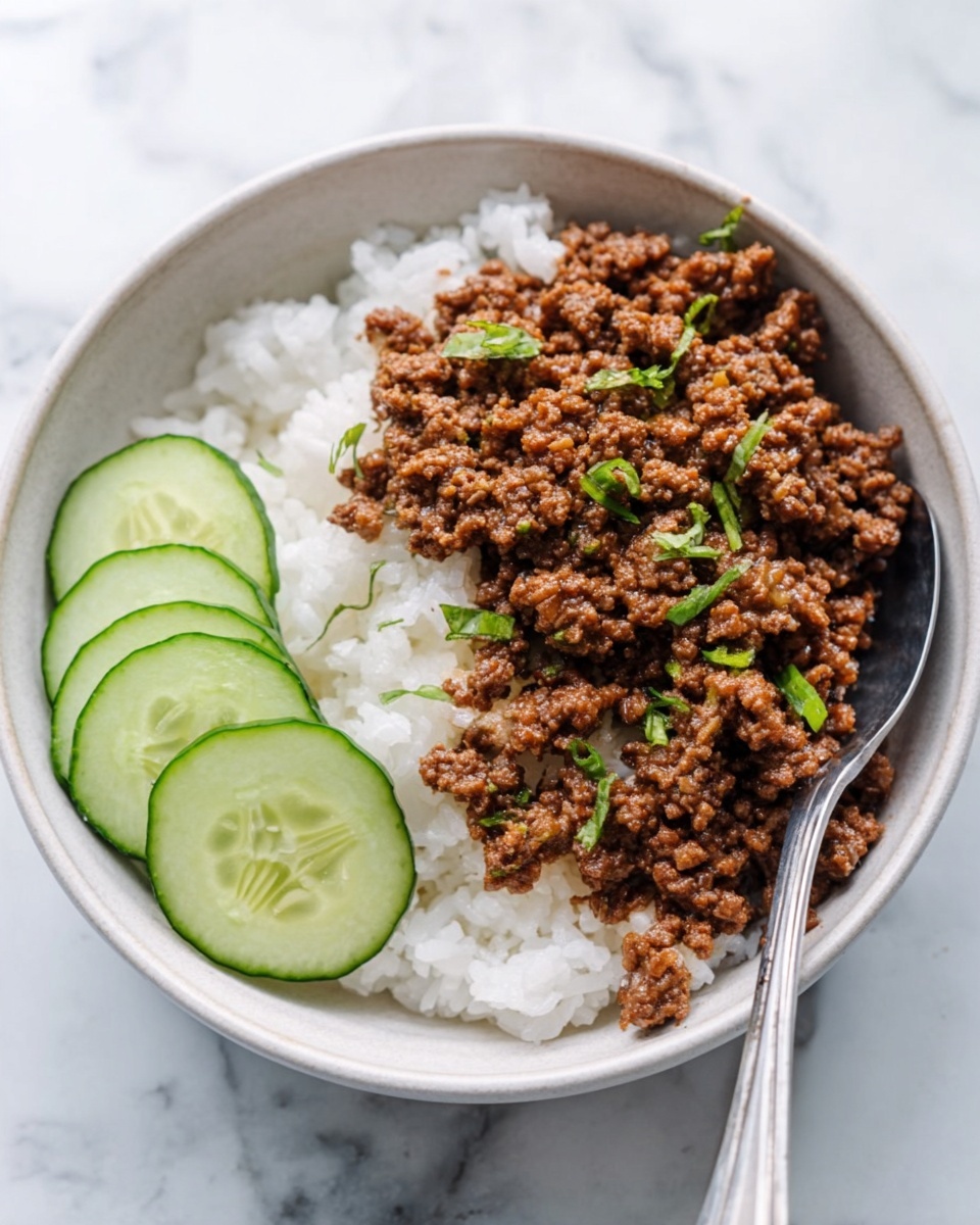A white bowl on a white marbled surface holds a dish with two main layers. The bottom layer is white rice, soft and fluffy. On top of the rice is a layer of cooked ground meat, brown and crumbly with small green herbs sprinkled for garnish. On the side of the meat, there are five thin slices of cucumber, light green with darker green edges, arranged neatly in a fan shape. A silver spoon is placed inside the bowl near the right side. photo taken with an iphone --ar 4:5 --v 7
