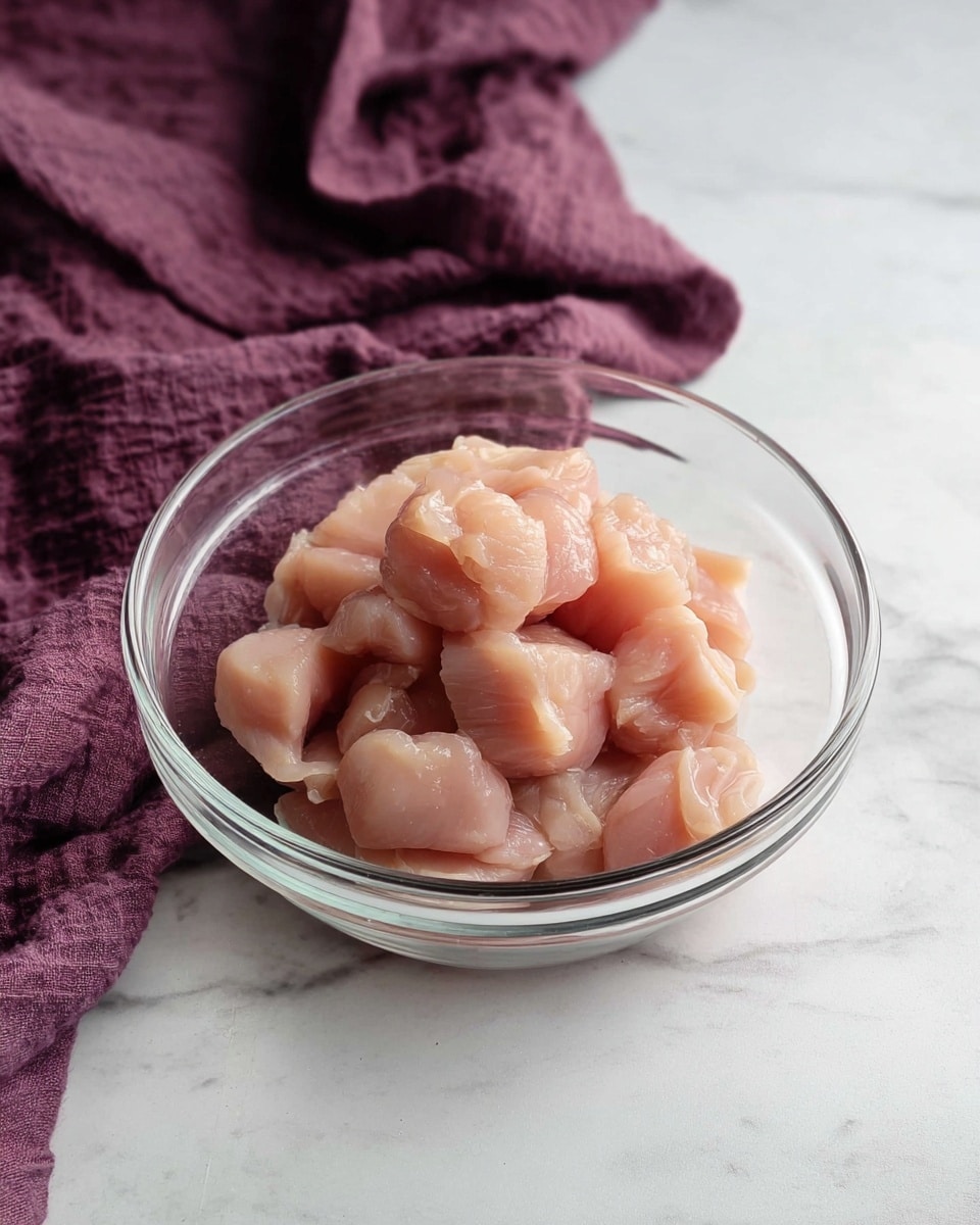A clear glass bowl filled with small, pale pink pieces of raw chicken placed at the center. The chicken pieces have a smooth, moist texture and are piled loosely in the bowl. The bowl sits on a white marbled surface, and behind it is a wrinkled dark purple cloth draped casually, adding a soft contrast to the scene. The overall setting is simple and clean, focusing on the chicken's natural color and shape photo taken with an iphone --ar 4:5 --v 7