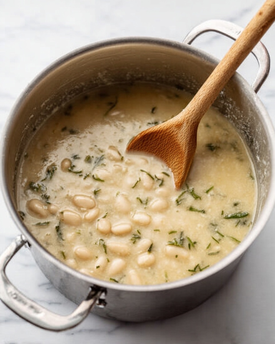 A silver metal pot filled with light creamy soup that has small white beans and green herb flakes mixed in, with a wooden spoon resting inside, the pot sitting on a white marbled surface photo taken with an iphone --ar 4:5 --v 7