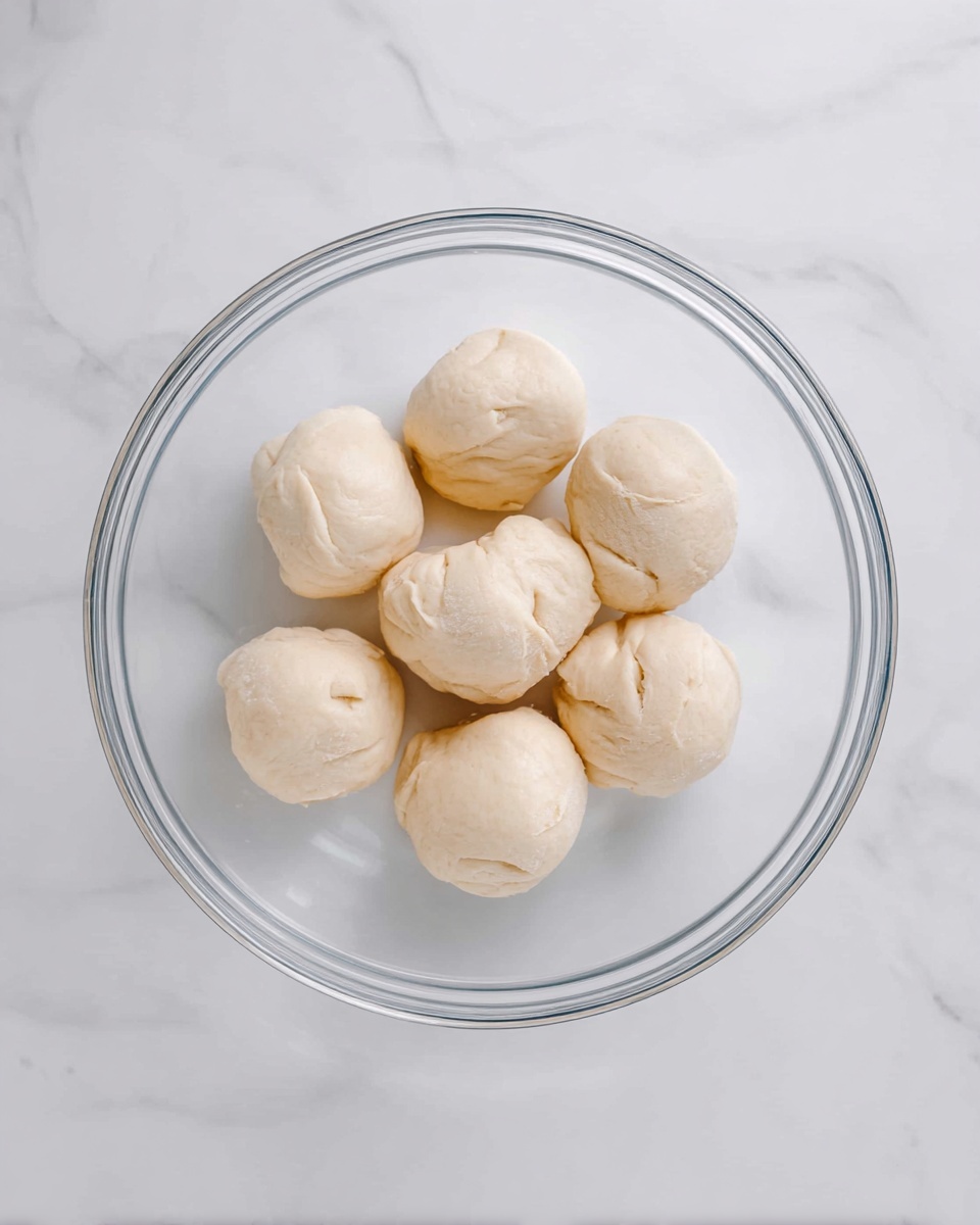 There are eight small, soft dough balls arranged in a circle inside a clear glass bowl. The dough balls are pale beige with some slight uneven texture, showing gentle folds and creases on their surfaces. The bowl sits on a white marbled texture, giving a clean and bright background. photo taken with an iphone --ar 4:5 --v 7