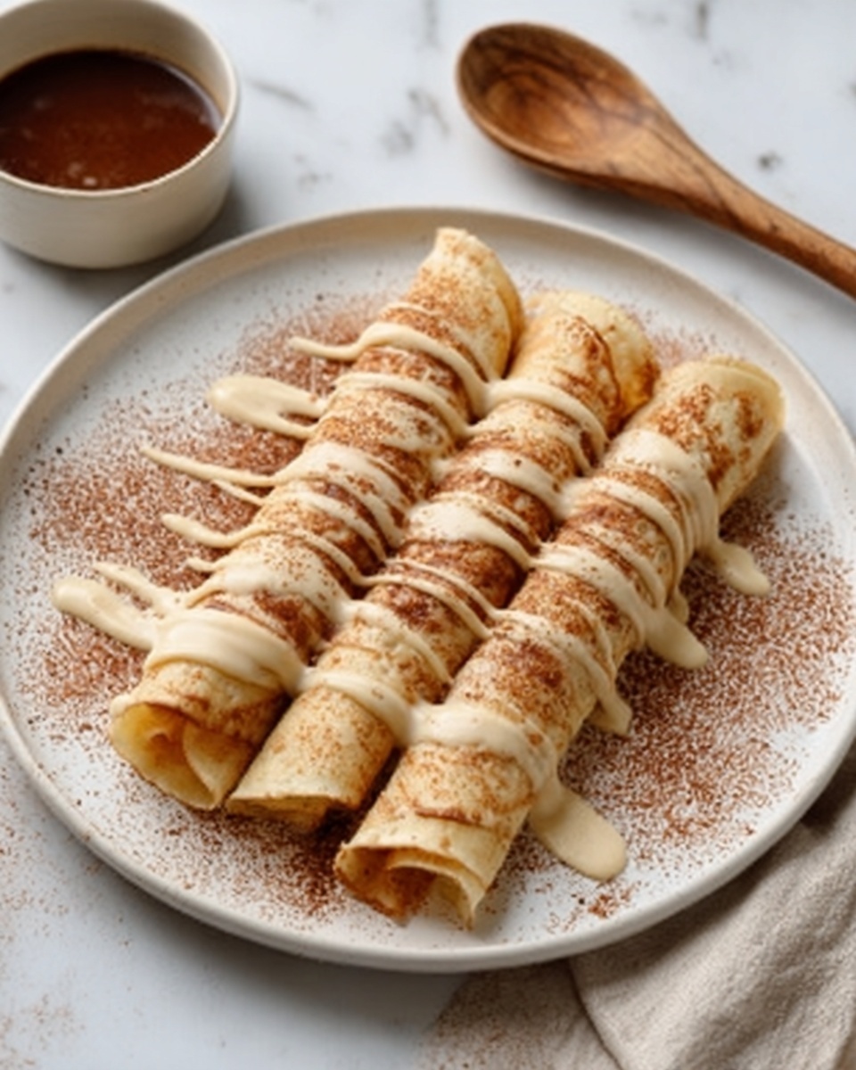 Three rolled light golden crepes are placed side by side on a white plate. The crepes are topped with a smooth creamy sauce drizzle running diagonally across each one. There is a light dusting of brown powder on the plate around the crepes. The plate sits on a white marbled surface, next to a wooden spoon and a small bowl with a dark brown sauce. Photo taken with an iphone --ar 4:5 --v 7