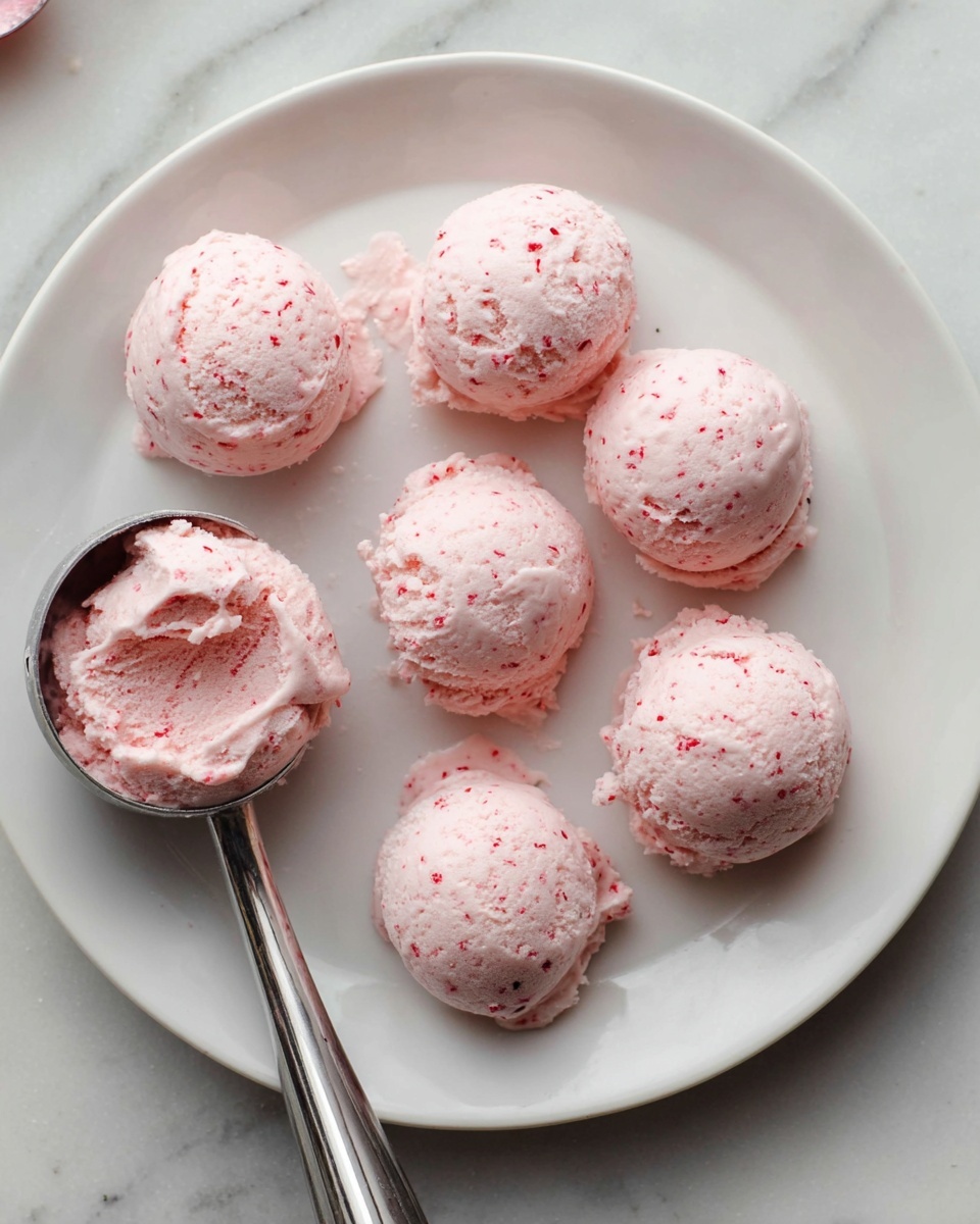 A white plate holds eight dollops of pink cream, each roughly round with a rough texture and small red specks spread throughout. The cream looks soft and slightly whipped, placed in two loose rows across the plate. A metal ice cream scoop rests on the edge of the plate, filled with the same pink cream, showing its smooth inside texture. The plate is on a white marbled surface with a faint pattern. Photo taken with an iphone --ar 4:5 --v 7
