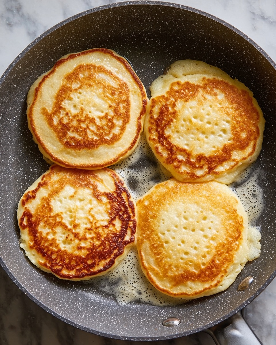 Four golden brown pancakes cook in a speckled gray frying pan. Each pancake shows small air bubbles on the surface with slightly crispy, darker golden edges. The pancakes are round but a bit uneven in shape, and light butter or oil bubbles around their edges in the pan. The gray pan contrasts with the warm, light brown color of the pancakes. The background is a white marbled texture. photo taken with an iphone --ar 4:5 --v 7