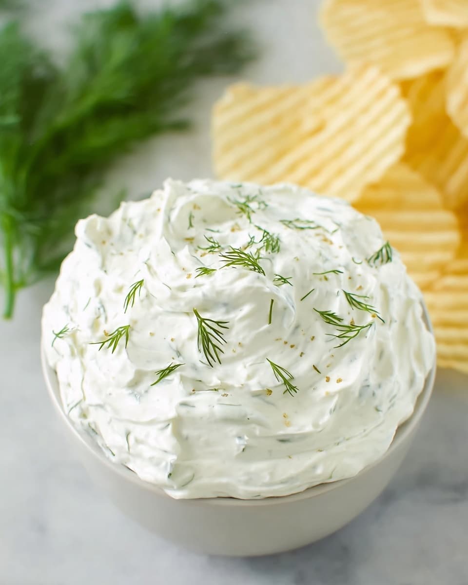 A white bowl filled with a thick layer of creamy white dip topped with small green dill sprigs scattered unevenly on the surface. The dip has a smooth but slightly textured look with soft peaks. The bowl sits on a white marbled surface, with fresh green dill leaves blurred in the background on the left and pale yellow ridged potato chips blurred on the right. Photo taken with an iphone --ar 4:5 --v 7