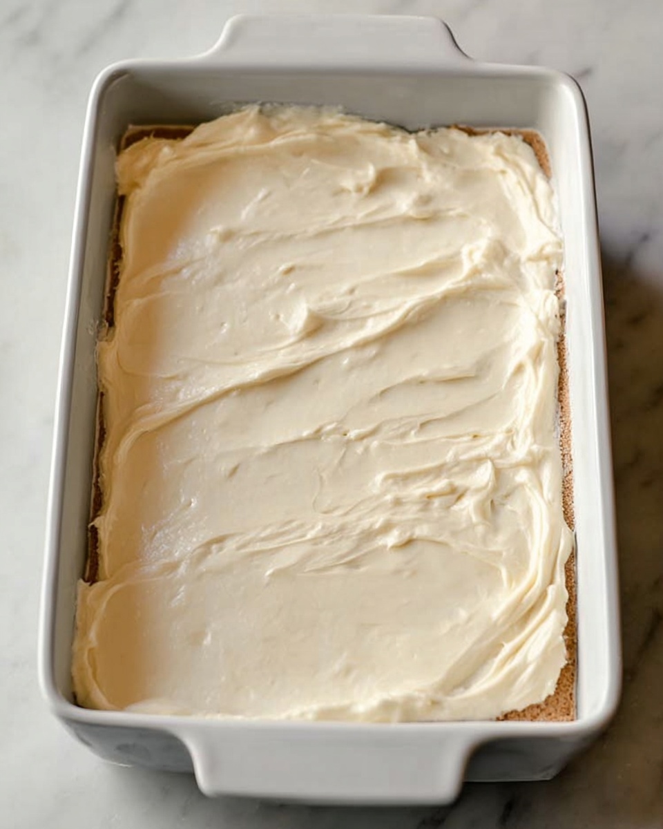 A white rectangular dish filled with two visible layers; the bottom layer is light brown with a soft texture, and the top layer is a thick, smooth cream-colored frosting spread evenly over the entire dish, with some gentle swirl patterns. The dish is placed on a white marbled surface photo taken with an iphone --ar 4:5 --v 7