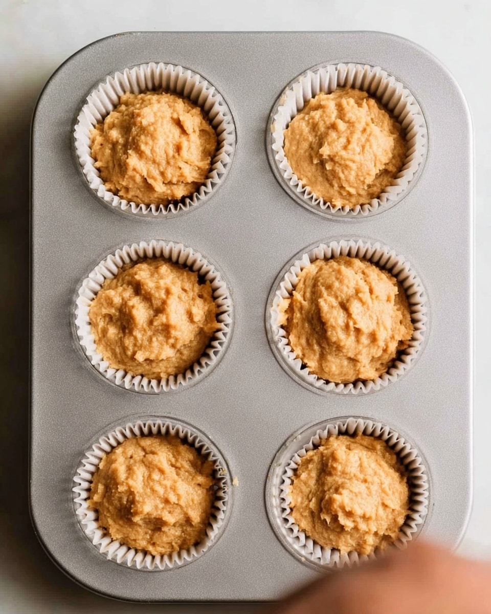A top-down view of six unbaked muffins placed in a silver metal muffin tray. Each muffin sits in white paper liners, showing a rough, slightly lumpy light brown batter. The muffins fill each liner unevenly, with some batter higher in the center. The tray is set on a white marbled surface, and a woman's hand is not visible in the frame. photo taken with an iphone --ar 4:5 --v 7