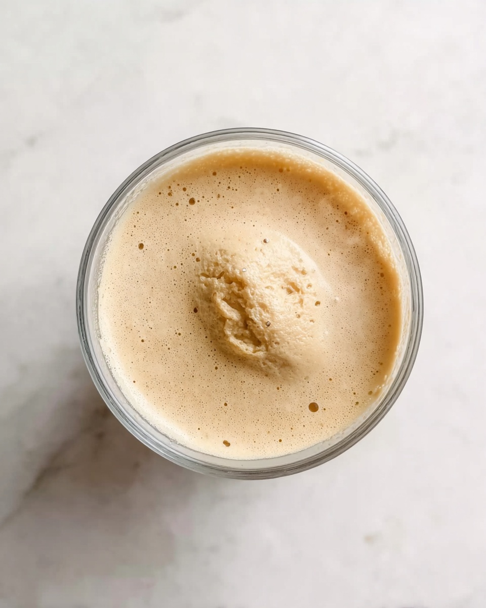 A clear glass cup filled with one layer of light tan foam, featuring a soft, uneven texture and a small mound in the center with slightly rough edges. The cup is placed on a white marbled surface, and the foam shows gentle bubbles and a creamy consistency. Photo taken with an iphone --ar 4:5 --v 7