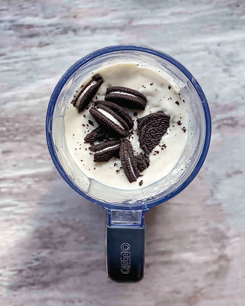 A clear round blender cup filled with a thick white creamy layer, topped in the center with several broken small black and white sandwich cookies. The blender cup sits on a white marbled surface with visible textured patterns. A dark blue handle extends downward from the cup, labeled with white text. Photo taken with an iphone --ar 4:5 --v 7