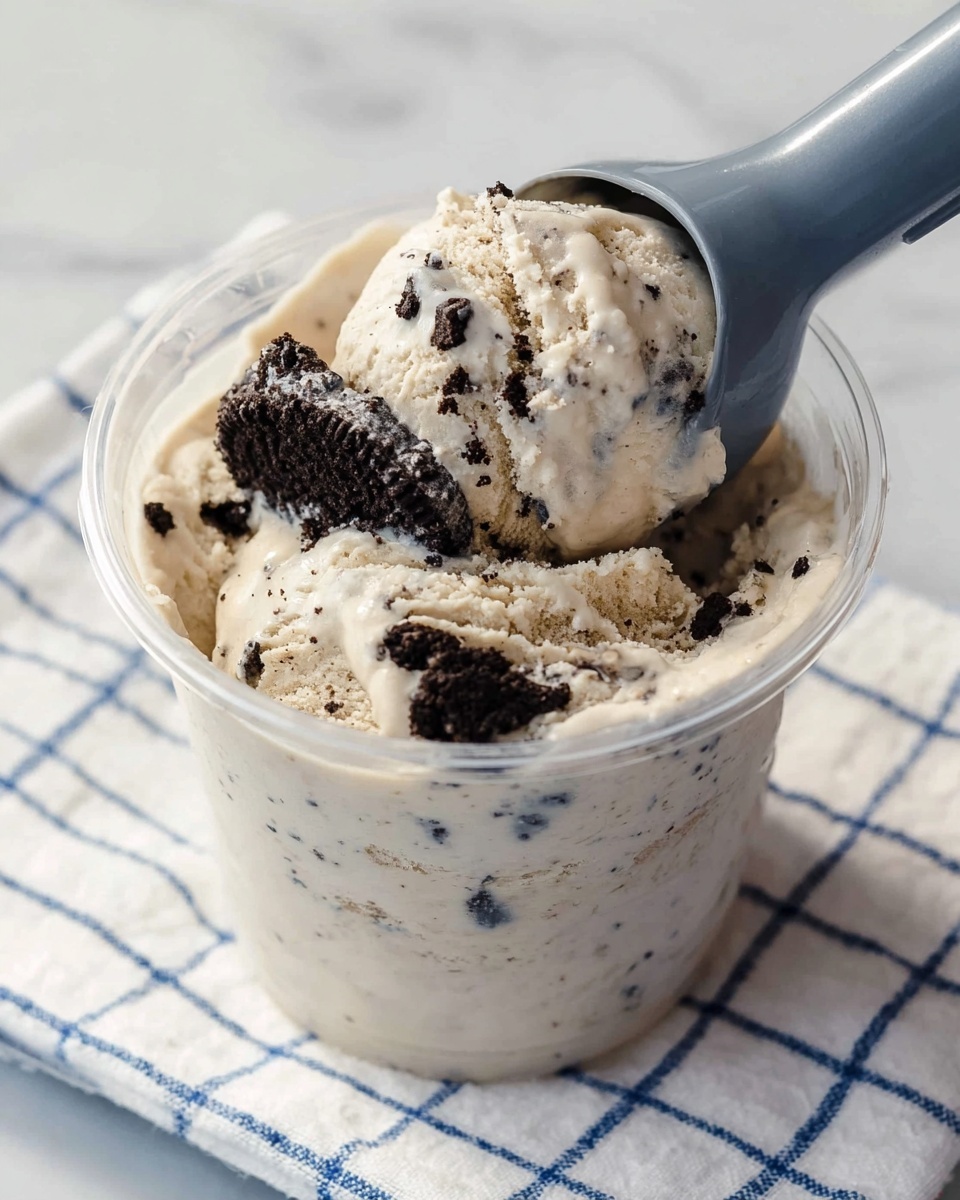 A clear plastic cup is filled with light beige ice cream that has small black cookie bits mixed evenly throughout. The ice cream is creamy and thick, with larger dark cookie chunks visible on top, one of which is being scooped by a gray ice cream scooper entering from the right side. The cup is placed on a white cloth with a blue grid pattern, set on a white marbled surface. The photo taken with an iphone --ar 4:5 --v 7
