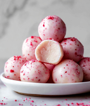 A pile of smooth, round, pale pink balls with tiny red specks scattered on their surface is stacked on a white plate. One ball is bitten to show a creamy, light pink inside texture. The balls are glossy and have a soft, velvety look, with some red specks sprinkled on and around the plate. The scene is set against a white marbled background. photo taken with an iphone --ar 4:5 --v 7