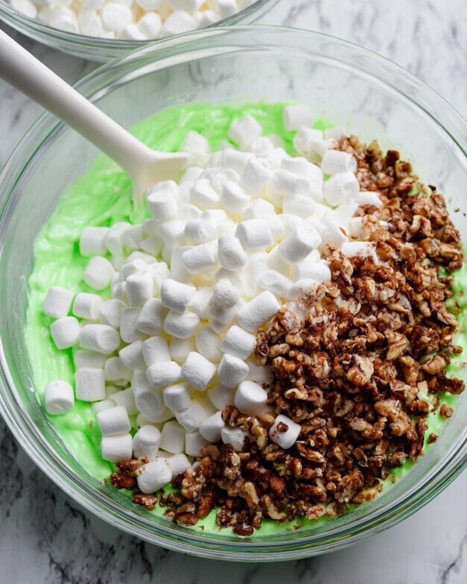 The image shows a clear glass bowl on a white marbled surface, filled with three main layers: the bottom layer is bright green melted marshmallow spread out thinly, the middle layer consists of a large pile of small, white mini marshmallows that look soft and fluffy, and the top layer is a heap of chopped brown nuts with a rough texture and uneven shapes. A white spoon rests on the right side inside the bowl, partially under the mini marshmallows. Photo taken with an iphone --ar 4:5 --v 7