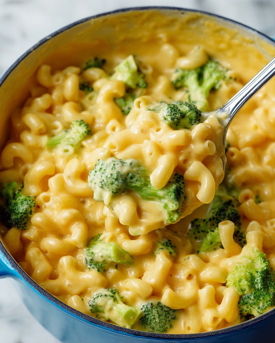 A close-up view of a creamy macaroni and cheese dish with bright green broccoli pieces mixed evenly throughout. The macaroni is smooth and coated in a thick, rich, golden-yellow cheese sauce. The pasta shapes are curved tubes, nestled among the small broccoli florets that add a fresh green contrast. A metal spoon is lifting a portion of the cheesy pasta, showing the thick texture of the sauce and the softness of the broccoli. The food sits inside a blue pot on a white marbled surface. photo taken with an iphone --ar 4:5 --v 7