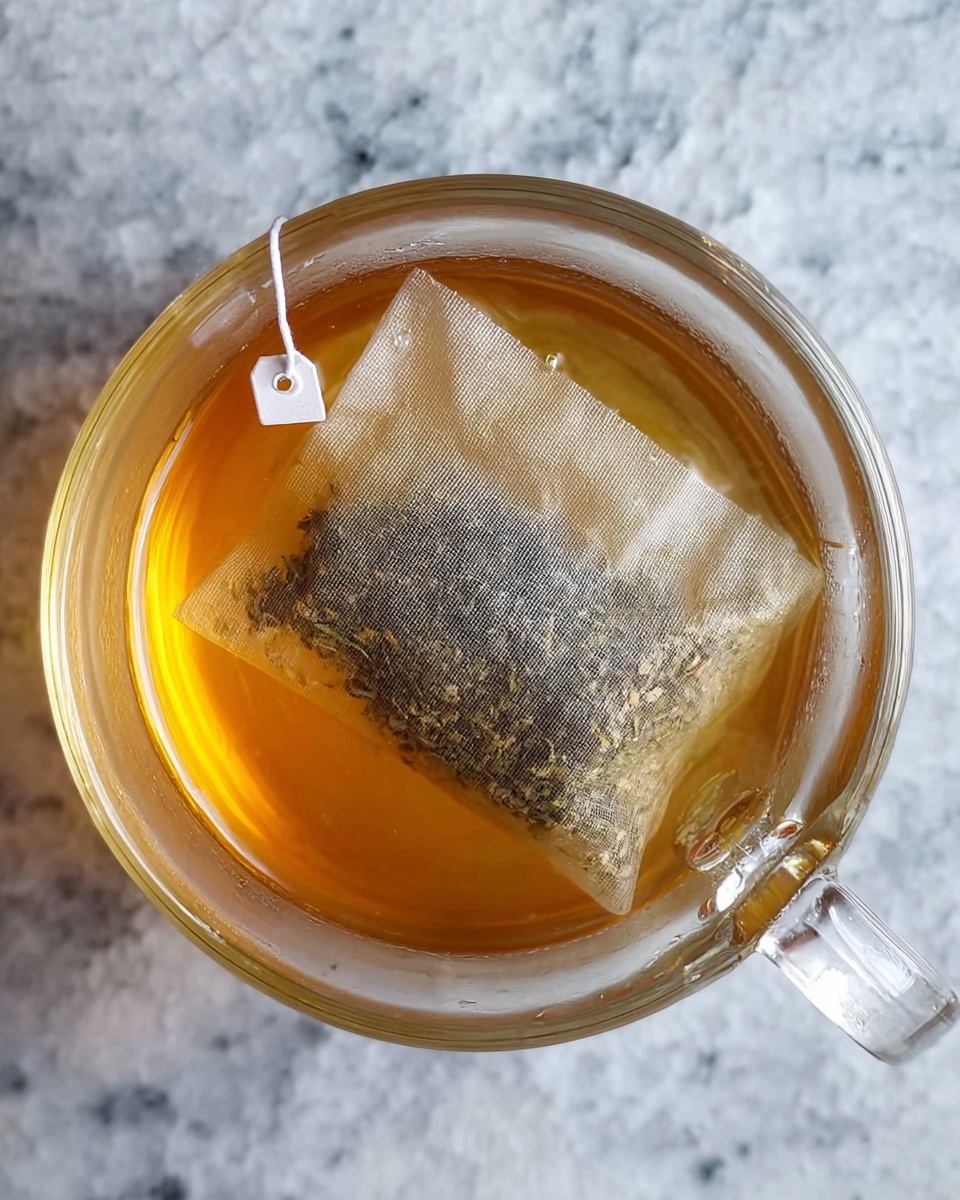 A clear glass cup shows light amber tea inside, with a tea bag fully soaked and resting at the bottom. The tea bag is rectangular and made of a fine mesh material, filled with loose tea leaves and small herbs visible inside. A white string with a small tag is attached to the tea bag, hanging over the edge of the glass. The cup is placed on a white marbled surface, and the photo is taken from a top-down view. photo taken with an iphone --ar 4:5 --v 7