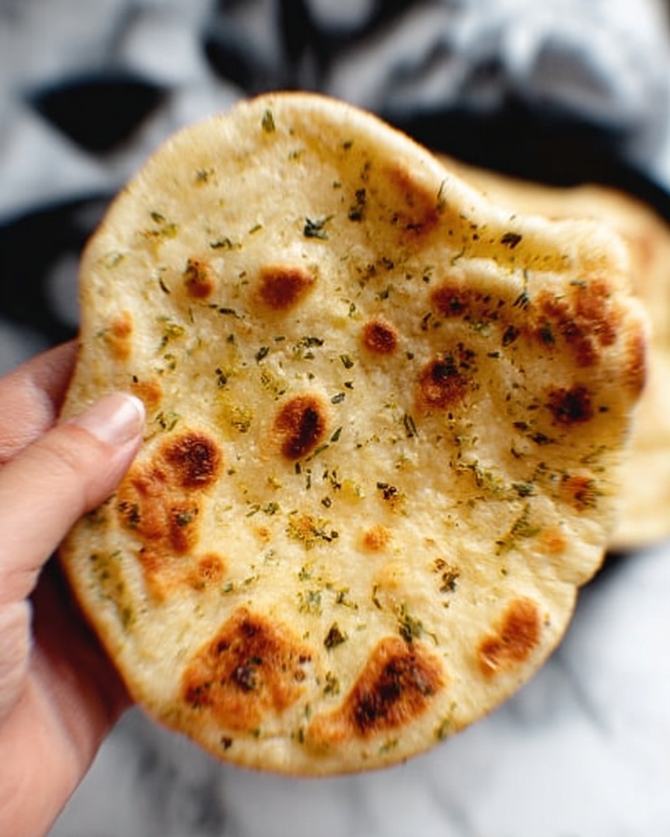 A woman's hand is holding a lightly browned flatbread with a soft texture, sprinkled with small pieces of herbs and bits of garlic, showing some gentle bubbles and a few toasted spots. The background is a soft white marbled surface with blurred dark shapes. photo taken with an iphone --ar 4:5 --v 7