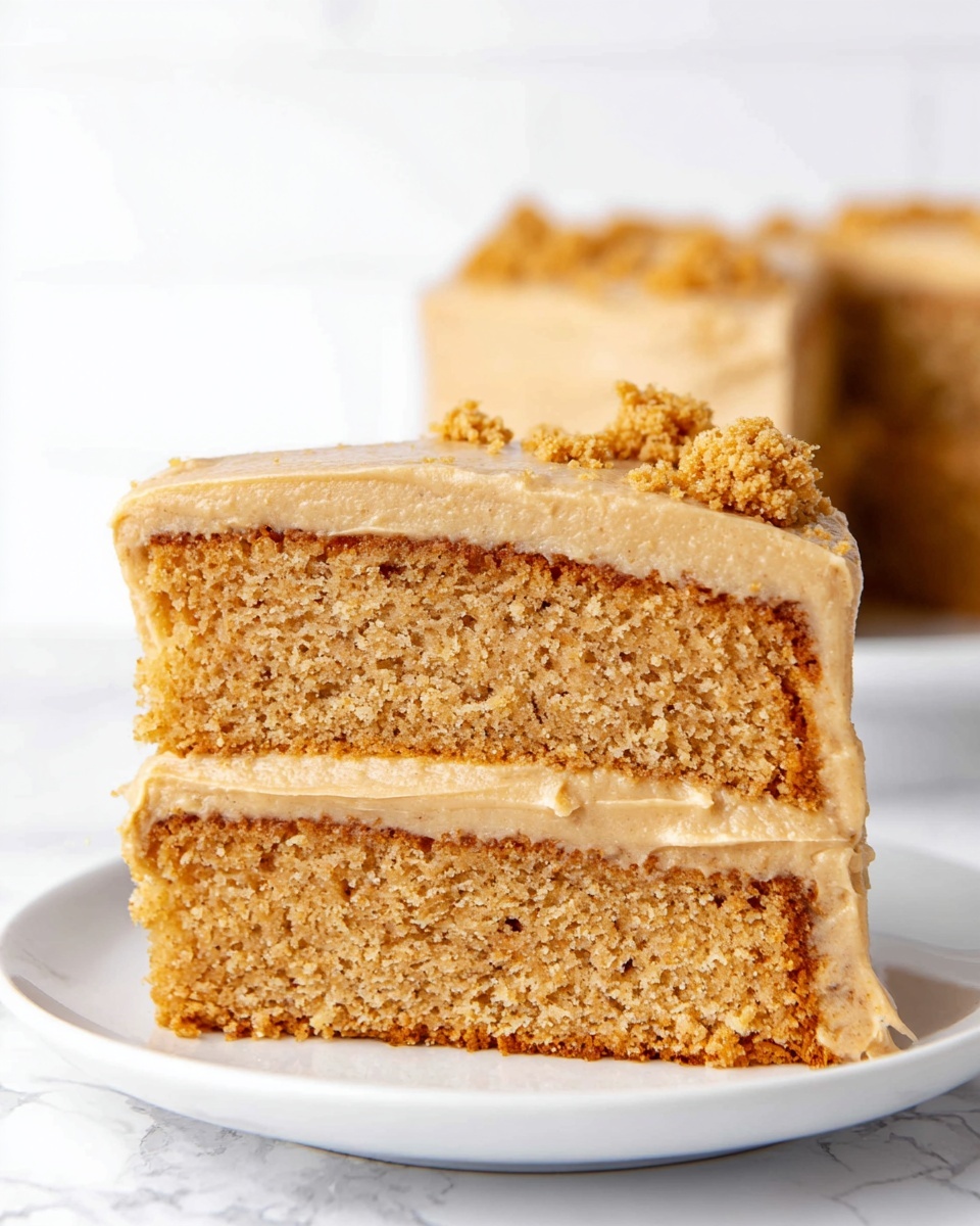 A slice of two-layer cake is shown on a white plate, set against a white marbled background. The cake has a light brown, moist texture with a creamy light brown frosting layer between the two cake layers. The top of the cake is covered with a smooth layer of the same light brown frosting, decorated with small crumbled pieces matching the cake color. The overall look is soft and inviting. Photo taken with an iphone --ar 4:5 --v 7