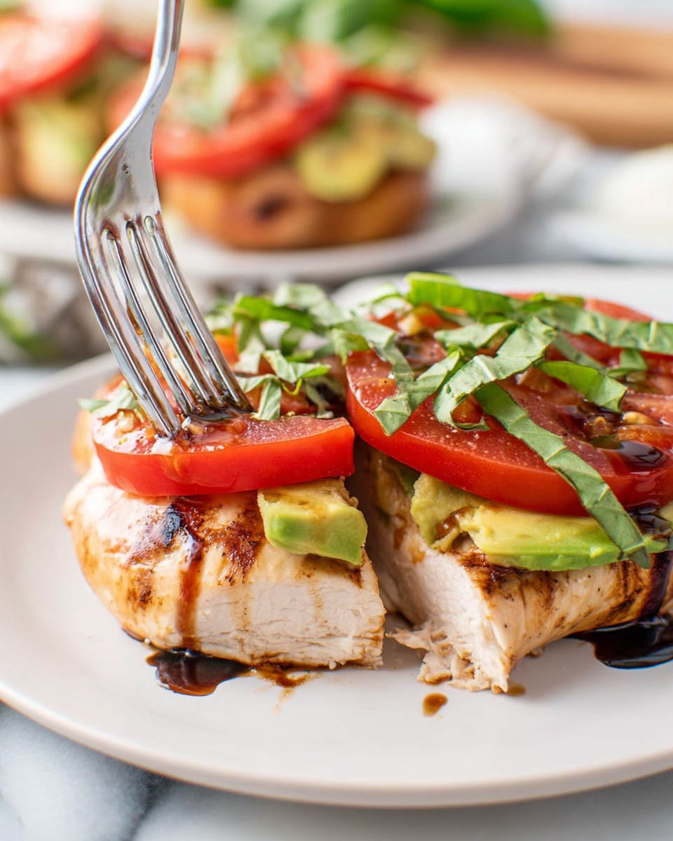 The image shows a white plate with a thick piece of cooked chicken at the bottom, light brown with grill marks. On top of the chicken, there is a layer of creamy avocado spread in green color, followed by a layer of bright red tomato slices. Thin strips of fresh green basil leaves are scattered on top, with a dark drizzle of balsamic glaze adding a shiny finish. A metal fork is gently pressing into the side of the chicken from the left side of the image. The background is a white marbled surface with a blurred view of another similar dish behind. Photo taken with an iphone --ar 4:5 --v 7
