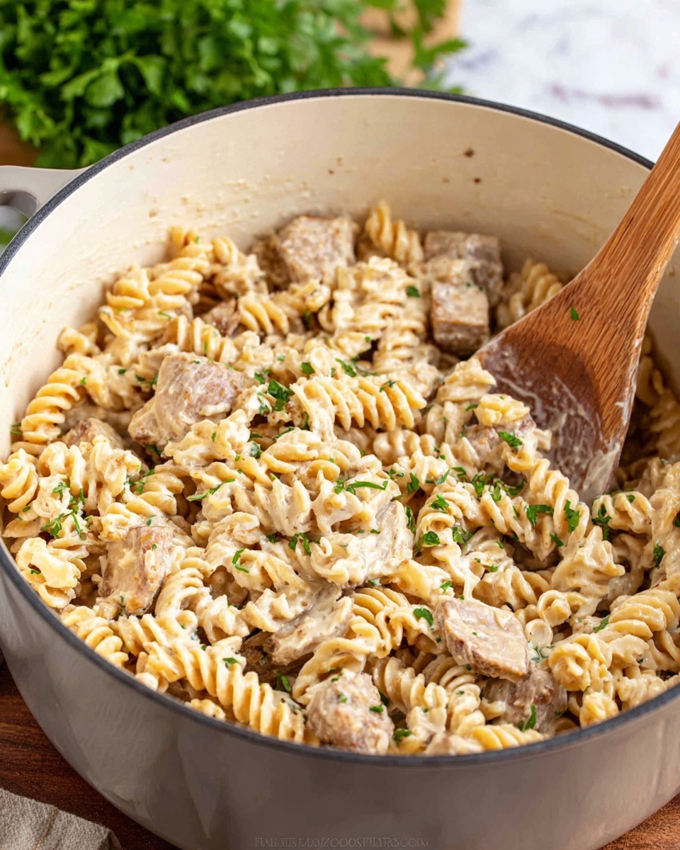 A large white pot filled with creamy pasta featuring spiral-shaped noodles coated in a rich, light beige sauce. Mixed in are thick strips of light brown cooked meat, and small pieces of green herbs scattered throughout add a touch of color. A wooden spoon rests inside the pot, partially submerged in the pasta. The background shows a blurred green herb bunch and a wooden surface beneath, replaced here with a white marbled texture. photo taken with an iphone --ar 4:5 --v 7