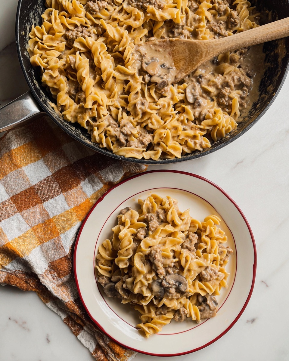 A white plate with a red line near the edge holds a creamy pasta dish made of short twisted noodles covered in a light brown sauce with small pieces of ground meat and sliced mushrooms mixed evenly throughout. Next to it is a black pan filled with more of the same pasta, showing the sauce glossily coating the noodles and meat, with a wooden spoon resting in the pan, partially covered in sauce. Below the pan and plate is a towel with a checkered pattern of white, beige, and orange, all placed on a white marbled surface. photo taken with an iphone --ar 4:5 --v 7