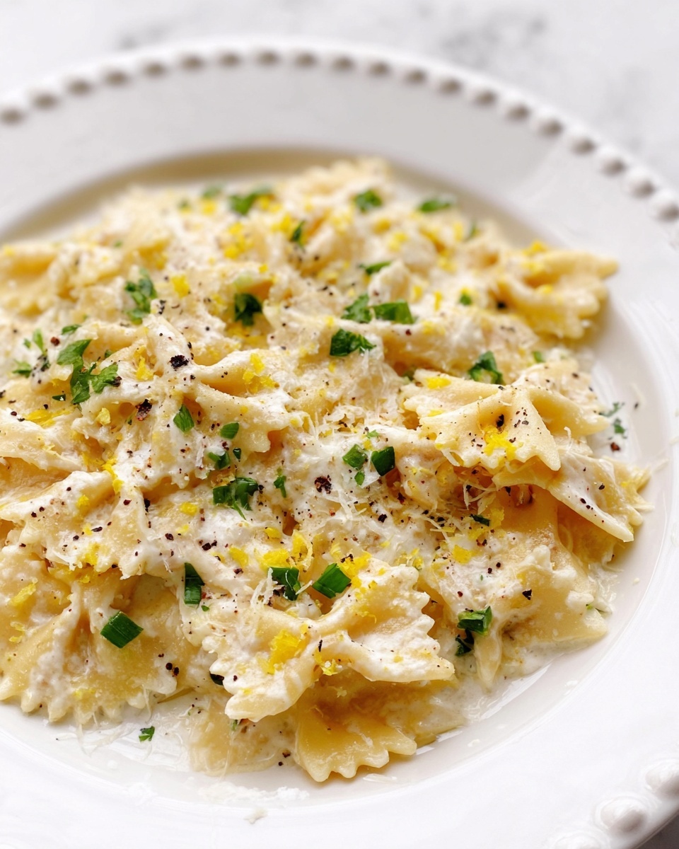 The dish is served on a white plate with a beaded edge, placed on a white marbled surface. It has about two layers of farfalle pasta mixed with a creamy white sauce, sprinkled generously with grated cheese. Small green herb pieces are scattered on top, adding contrast to the pale yellow pasta and white sauce. There are also tiny black pepper flakes and bits of yellow zest sprinkled on the dish, giving a fresh and textured look. The overall presentation is close-up, showing the creamy sauce and toppings clearly. photo taken with an iphone --ar 4:5 --v 7