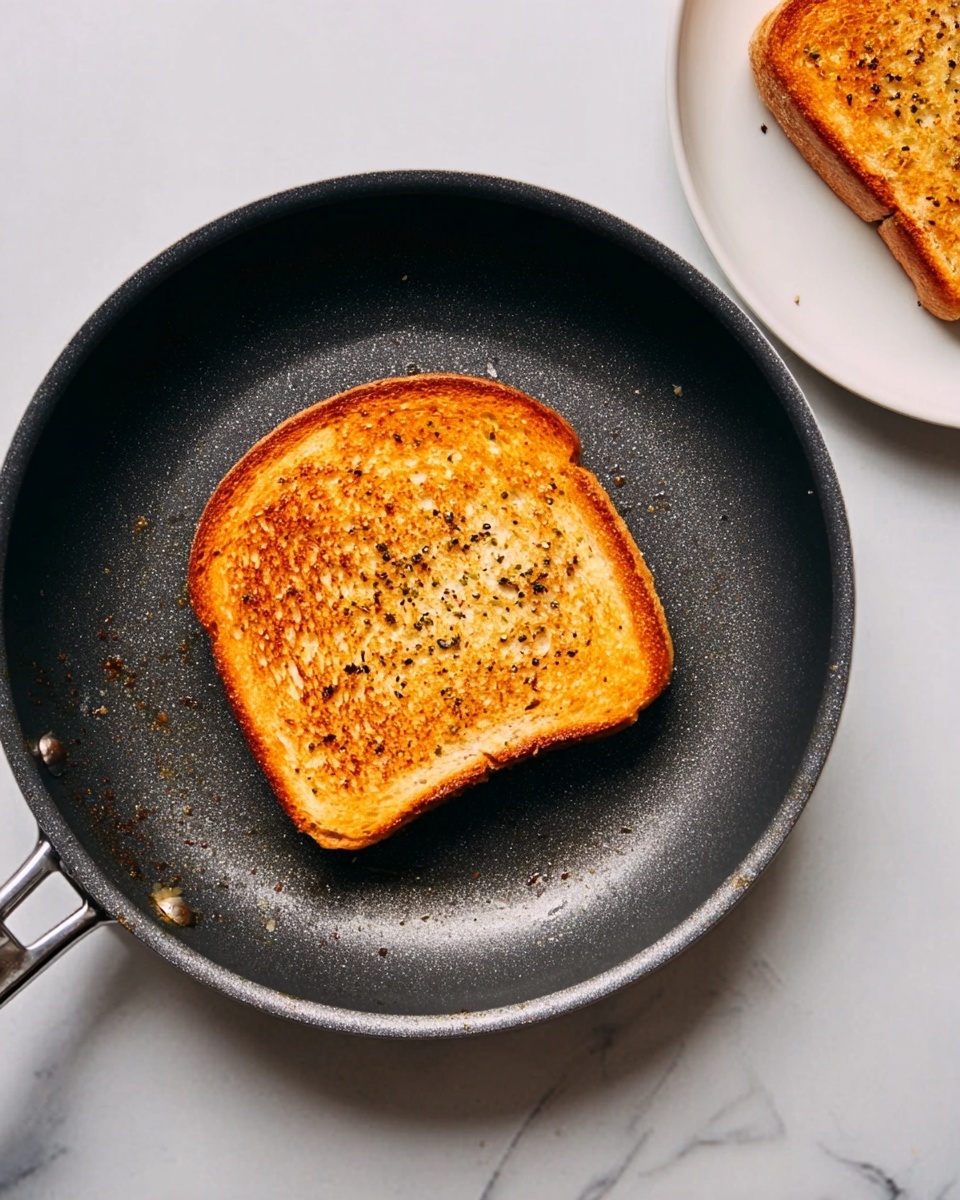 A single golden-brown slice of toast with specks of black seasoning sits in the center of a dark frying pan, showing a crunchy texture on top. The frying pan is positioned on a white marbled surface, and nearby to the right, there is a white plate with a matching golden-brown toasted slice. The overall colors highlight warm tones of toasted bread against the dark pan and bright white background. Photo taken with an iphone --ar 4:5 --v 7