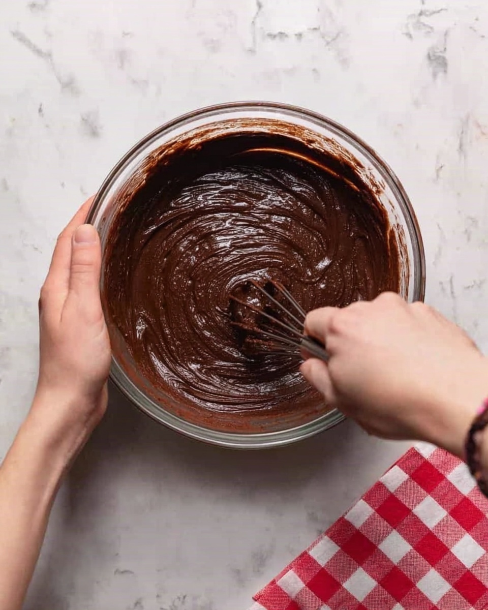 A clear glass bowl filled with dark brown chocolate batter is shown from above, with a woman's hand holding the bowl steady on the left side while the other woman's hand is stirring the batter with a whisk, creating smooth, swirling textures on the surface. The bowl is placed on a white marbled surface with a red and white checkered cloth partially visible on the right. photo taken with an iphone --ar 4:5 --v 7
