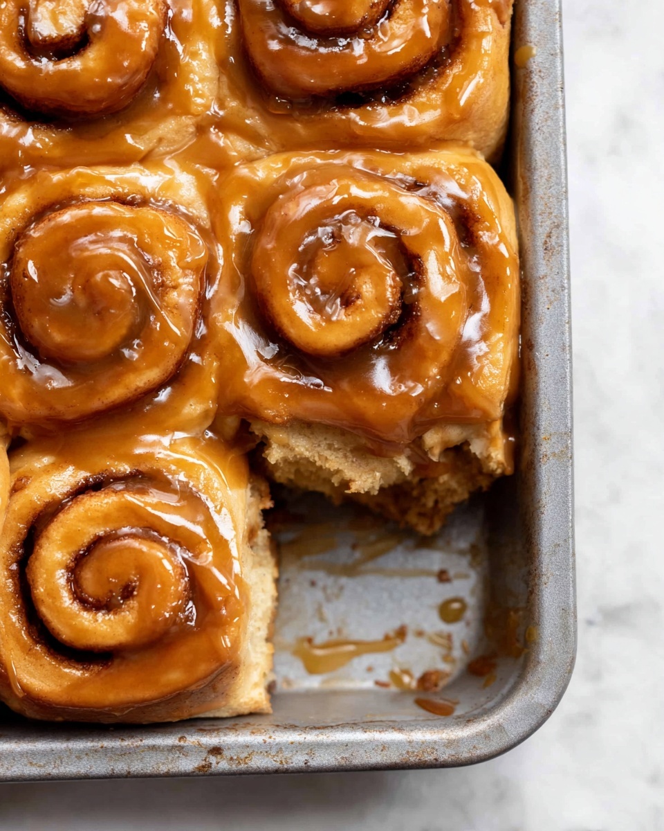 The image shows a close-up of four cinnamon rolls in a metal baking pan, arranged in a square shape with one roll separated from the rest. Each cinnamon roll has two visible layers: the soft, light golden brown dough base with a tight spiral, and a thick, glossy caramel-colored glaze generously covering the top, creating a shiny texture. The rolls have a slightly sticky and smooth surface, with the spiral revealing some dark brown cinnamon filling inside. The metal pan has a matte, slightly worn silver look with some caramel glaze drips around the rolls. The background is a white marbled surface. Photo taken with an iphone --ar 4:5 --v 7
