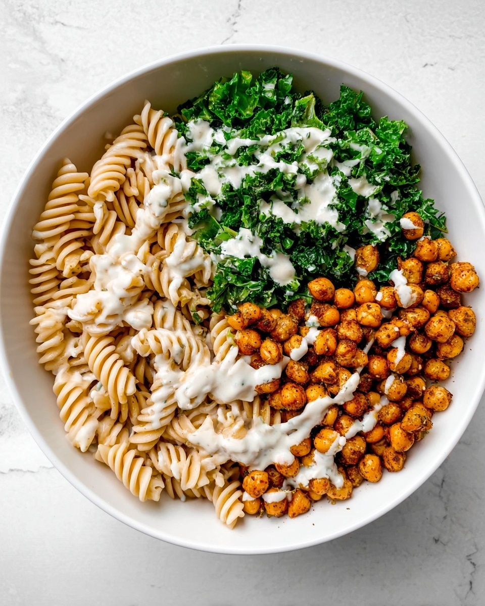 A white bowl is filled with three main sections: light beige spiral pasta on the left, bright green chopped kale on the top right, and golden-brown roasted chickpeas on the bottom right. All three parts are topped with a drizzle of white creamy sauce. The bowl sits on a white marbled surface with soft lighting. photo taken with an iphone --ar 4:5 --v 7