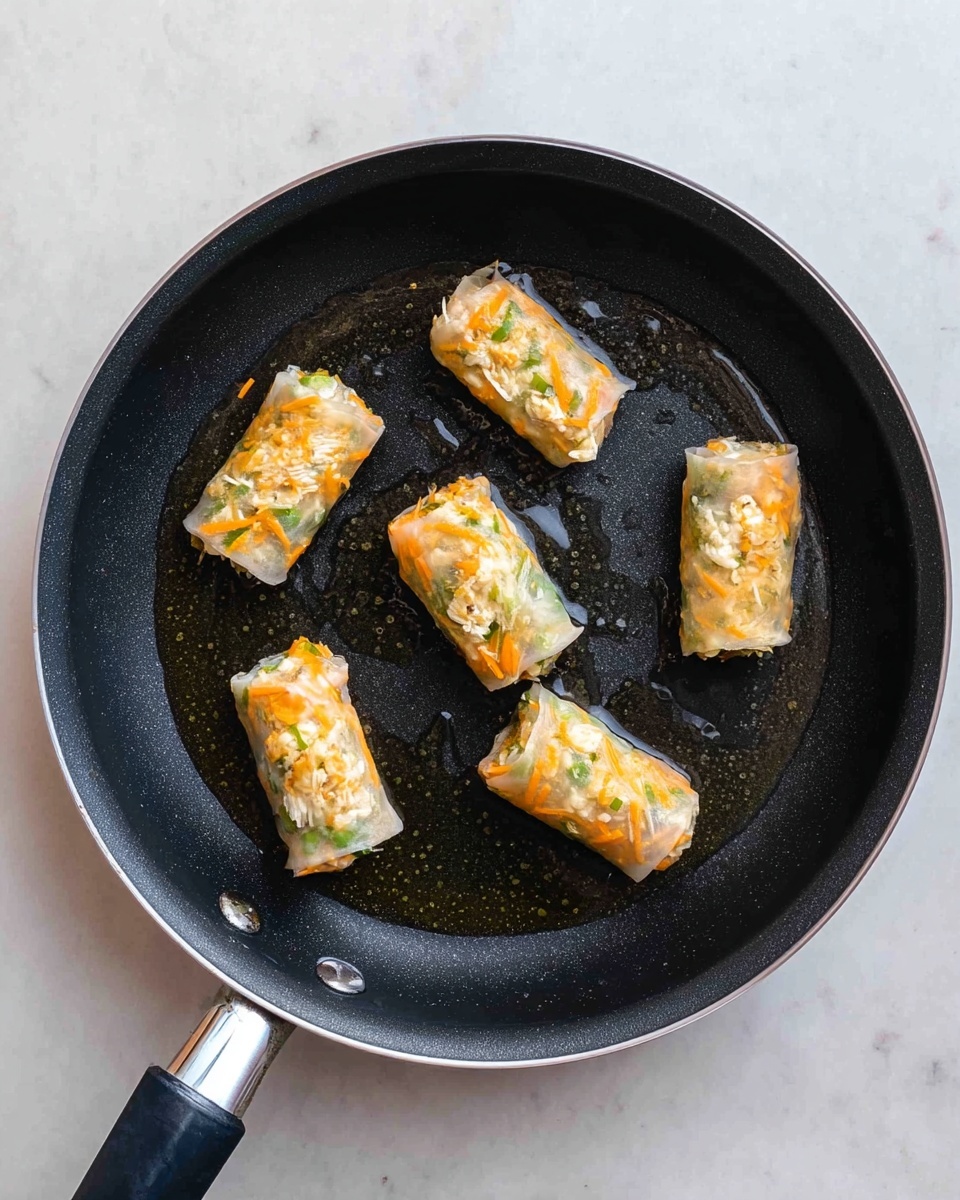There is a black frying pan with a silver edge and black handle placed on a white marbled surface. Inside the pan, five small rolls are being cooked. Each roll has a light, translucent outer layer that shows the filling inside, which is made of shredded orange carrot mixed with small green leafy pieces and crumbly white bits. The rolls are arranged loosely, with some oil glistening around them on the pan’s surface. photo taken with an iphone --ar 4:5 --v 7