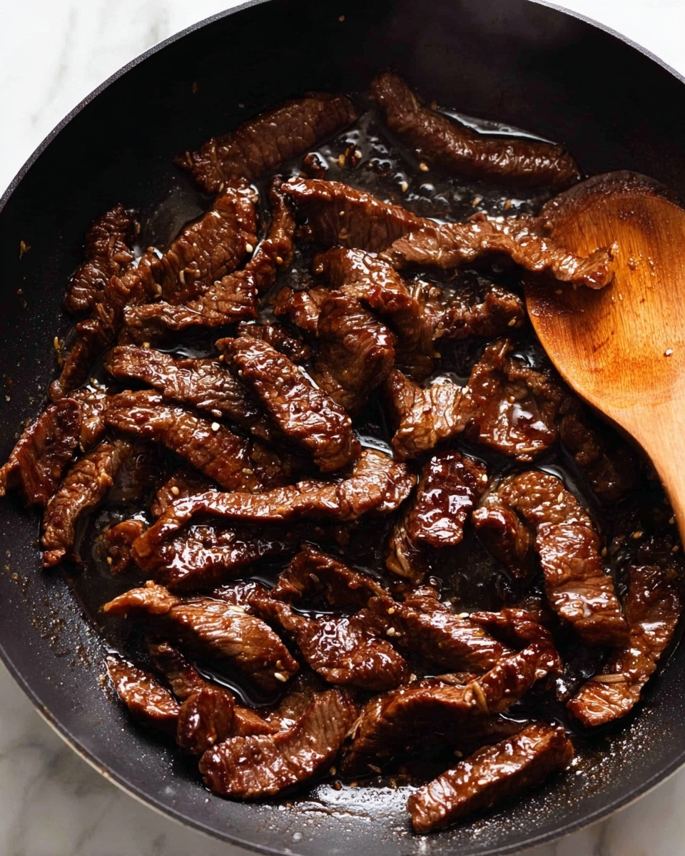 The image shows a black pan filled with cooked beef strips that are brown and glossy from being cooked in oil or sauce. The beef pieces are irregular in shape and cover the whole pan bottom. There is a wooden spoon resting on the edge of the pan, partly touching the beef, with a warm light wood tone and smooth texture. The background surface is white with a marbled pattern. photo taken with an iphone --ar 4:5 --v 7