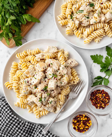 Two white plates sit on a white marbled surface, each filled with creamy rotini pasta mixed with chunks of light beige chicken pieces, all coated in a thick white sauce speckled with black pepper and green herbs. The pasta is topped with small green herb pieces and light shreds of cheese. A silver fork rests on the right side of the larger plate. To the side, a small clear bowl holds bright red chili flakes with yellow seeds. A wooden cutting board with fresh parsley leaves partially enters the top left corner while a black and white checkered cloth lies near the bottom left. The image is bright and clean, focused on the food. Photo taken with an iphone --ar 4:5 --v 7